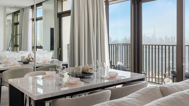 A modern dining area with a table set for two, large windows with curtains, and a view of a city skyline. - Serene 1BR Escape with Burj Khalifa View (Dubai)