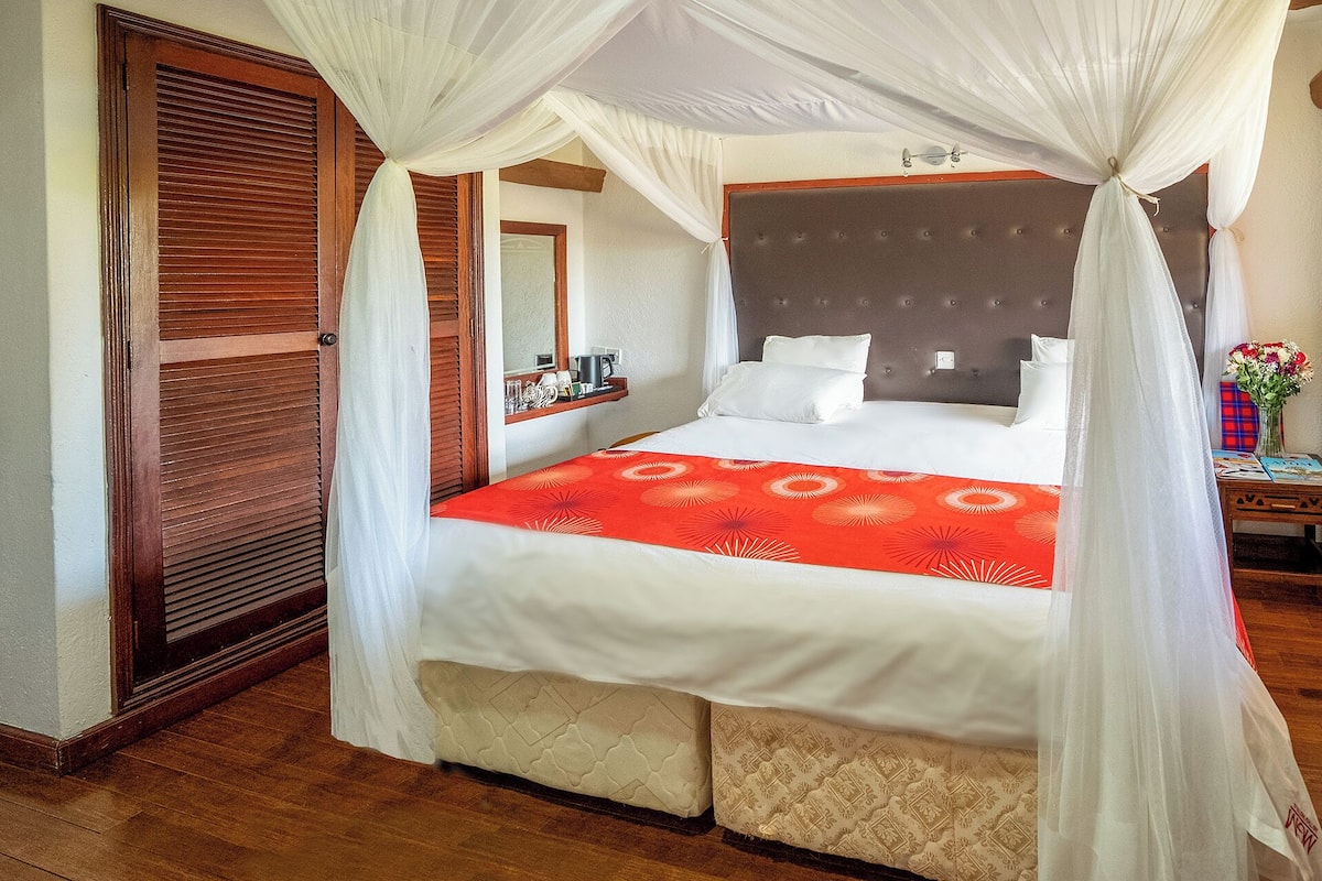 A bedroom with a four-poster bed, a wooden door, a bedside table with a vase, and a mirror.
