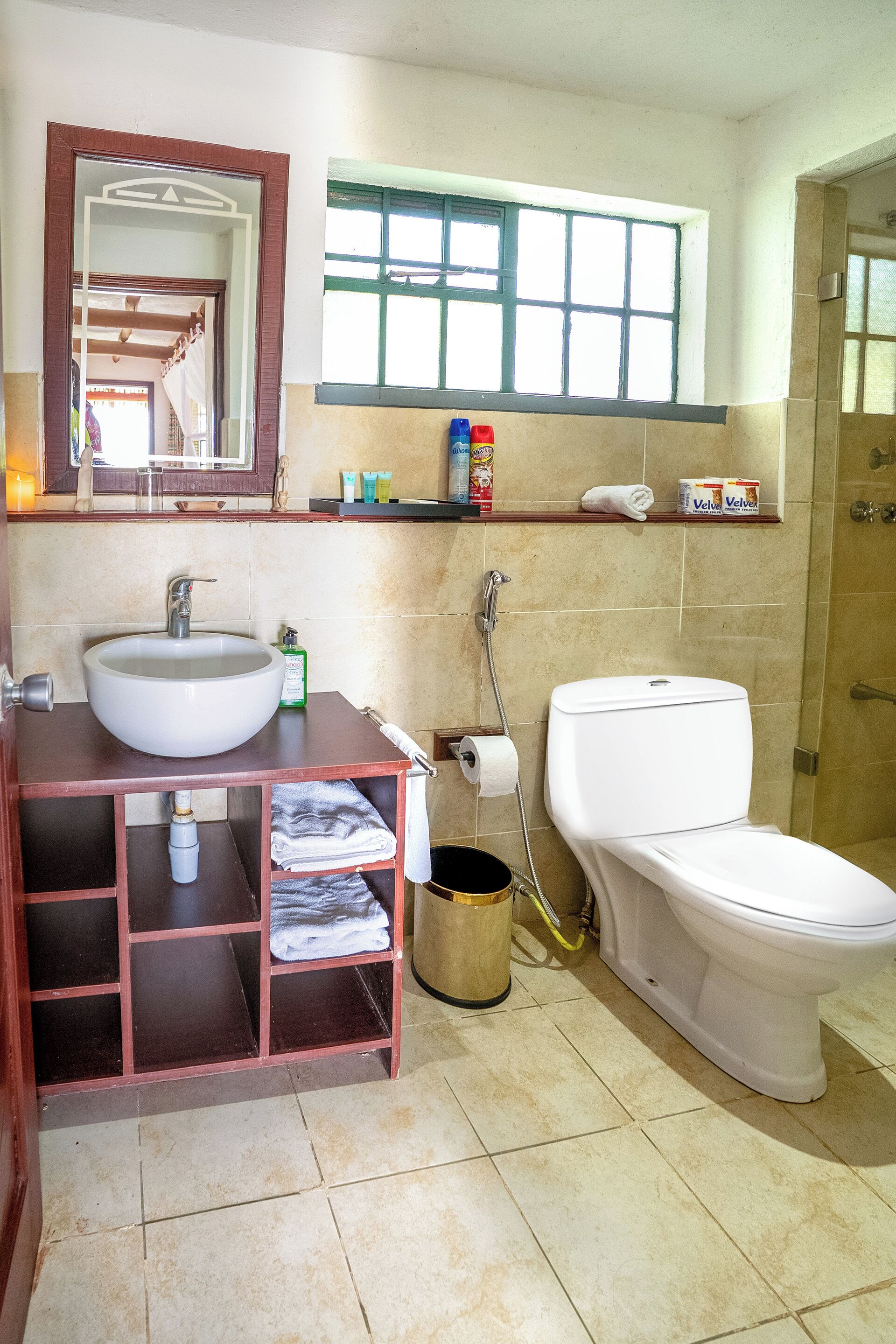 A bathroom with a sink, toilet, and shower. There is a mirror above the sink, a window above the toilet, and a shelf with toiletries.