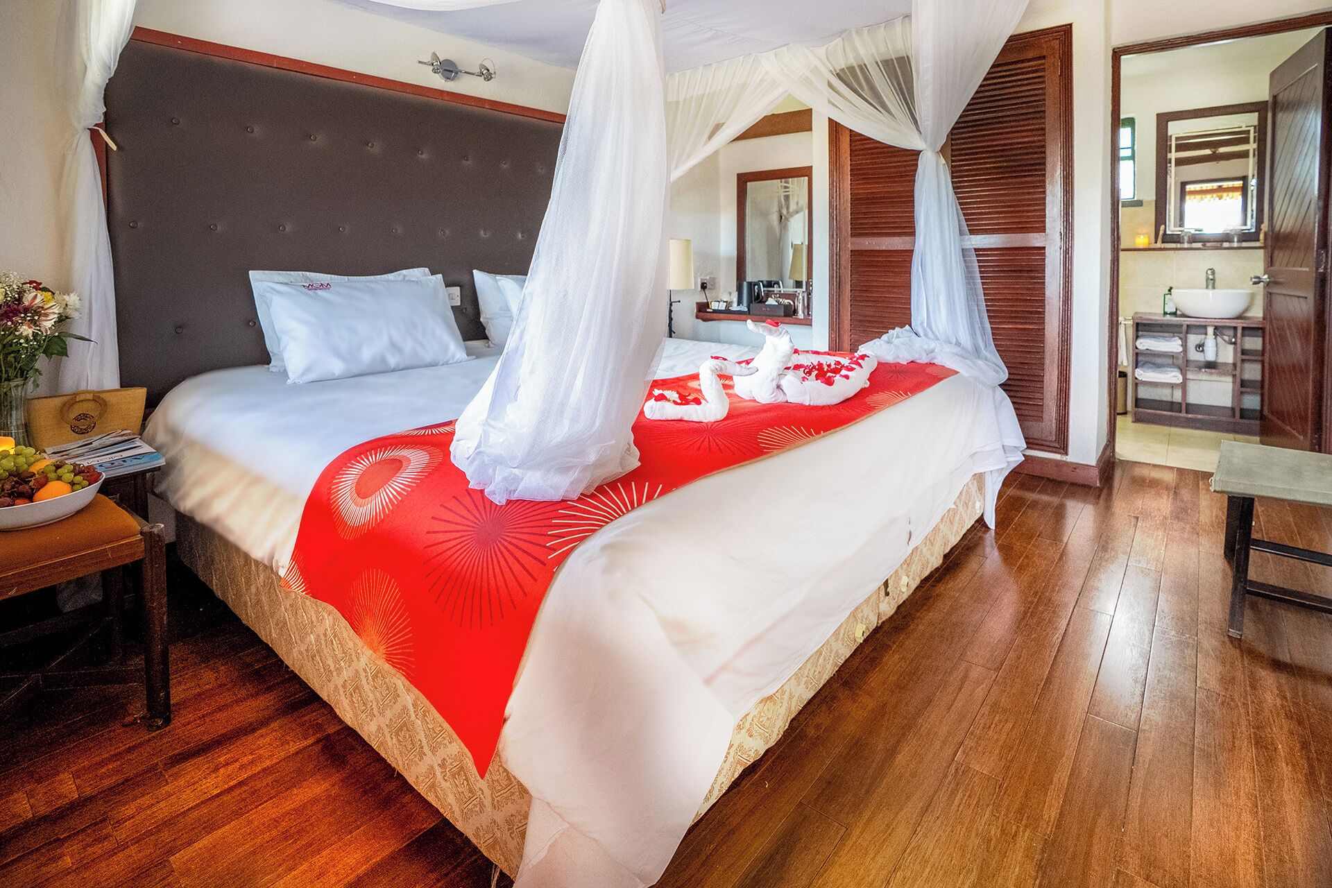 A four-poster bed with a red and white bedspread, a bedside table with a bowl of fruit, and a wooden floor.