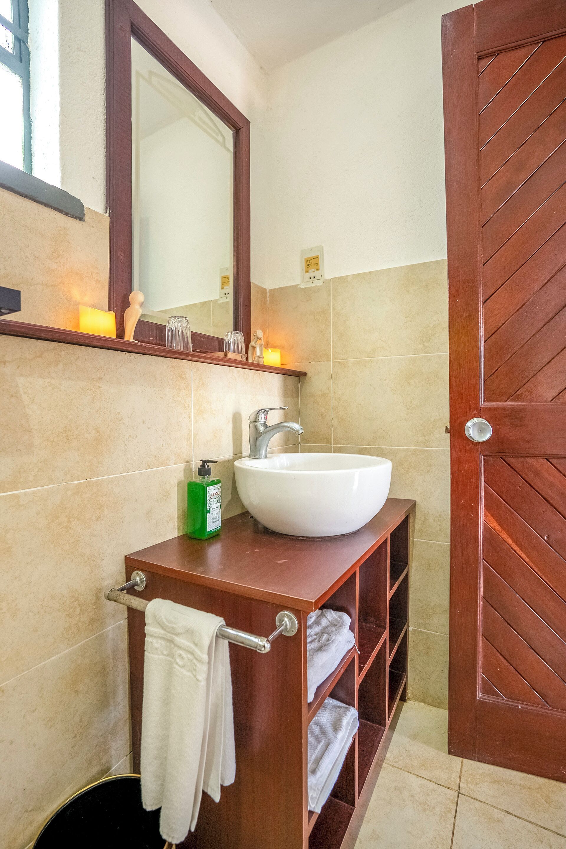 A bathroom with a wooden vanity, a white sink, and a mirror above it. There are towels hanging on a rack and a green bottle on the counter.