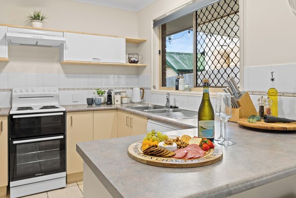 Una cocina moderna con armarios blancos, horno empotrado, encimera con una tabla de frutas y quesos, y una botella de vino.