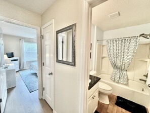A hotel room with a hallway leading to a bathroom. The bathroom has a white sink, a toilet, and a shower with a patterned curtain. A large mirror with an ornate frame is mounted on the wall above the sink. - Sweet Retreat - 3/2 w/ Golf Cart (Buttonwood, The Villages)