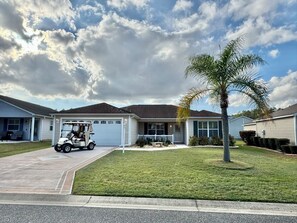 A single-story house with a garage, a lawn, and a palm tree. - Sweet Retreat - 3/2 w/ Golf Cart (Buttonwood, The Villages)