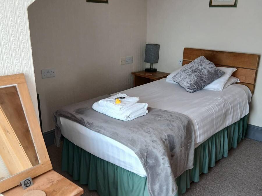 Ett hotellrum med en säng, ett nattduksbord, en lampa, en inramad tavla på väggen och ett skrivbord med en spegel.