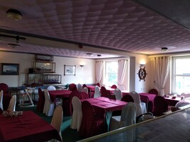 Uma sala de jantar com mesas postas para refeição, cadeiras com apoio para a cabeça, um leme na parede e quadros emoldurados.