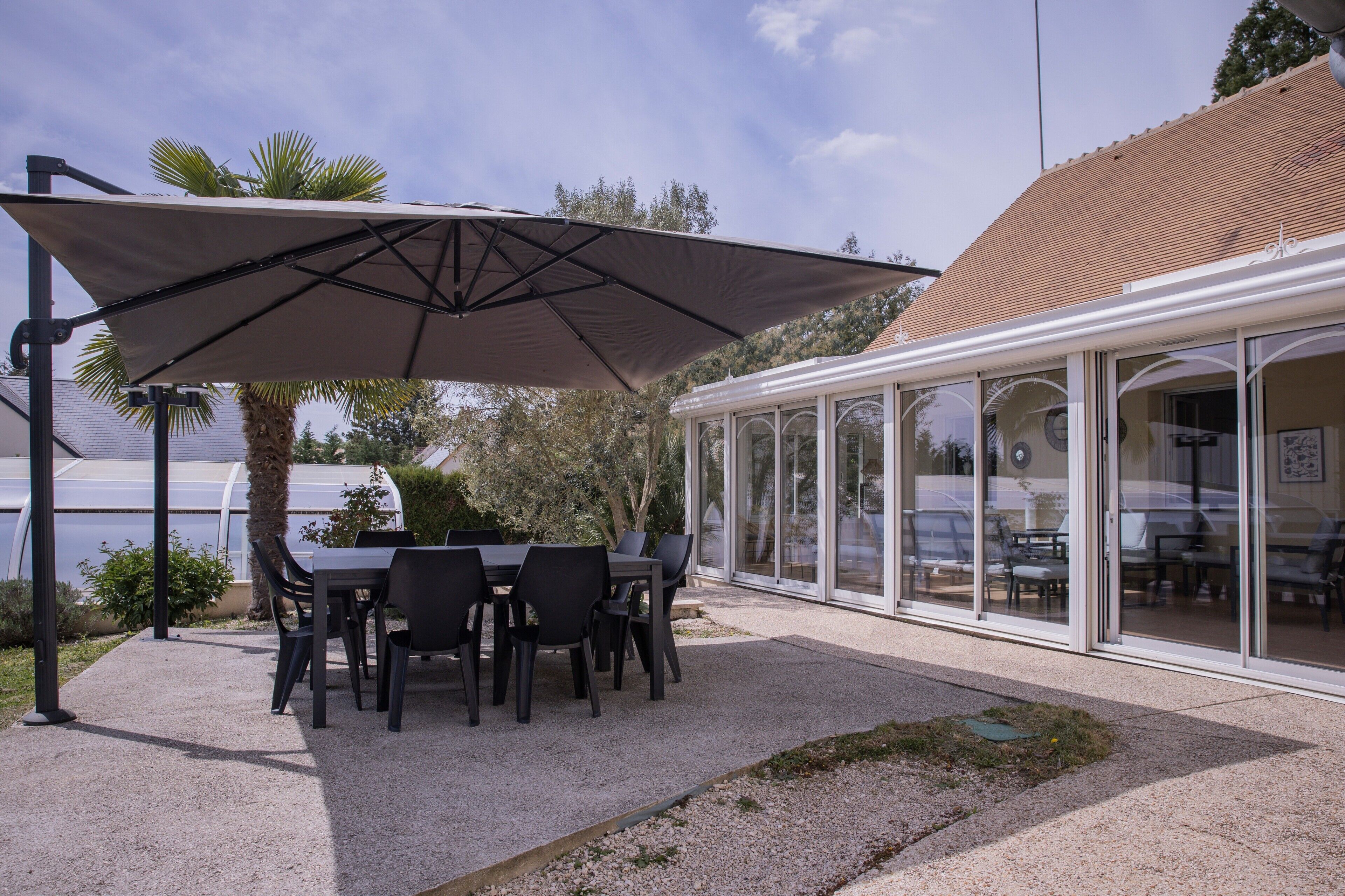Pátio externo com mesa e cadeiras, um grande guarda-sol e uma estufa com portas de vidro.
