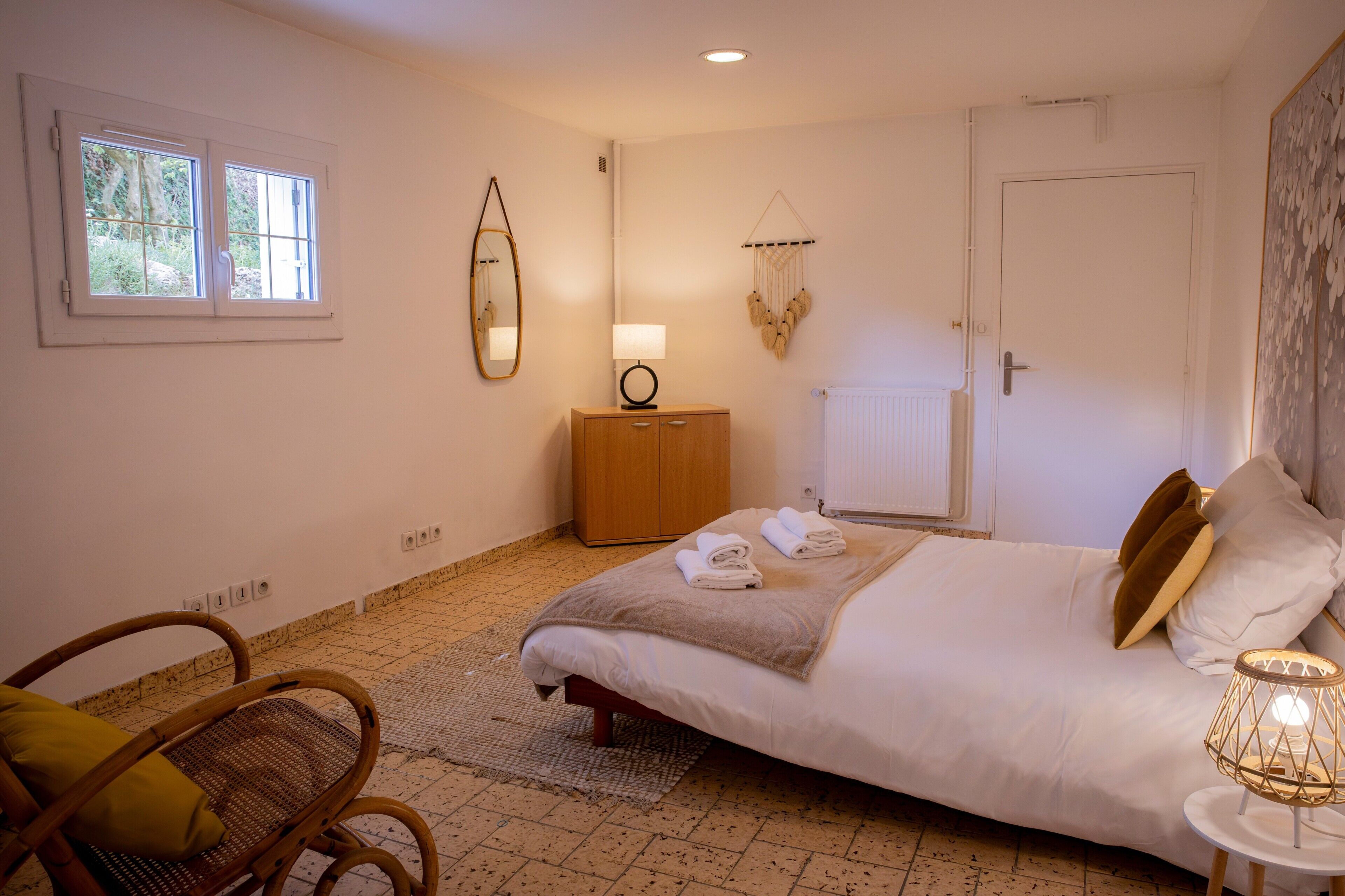 A bedroom with a bed, a bedside table, a chair, a mirror, a wall-mounted lamp, and a window.