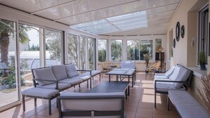 Um pátio ensolarado com teto de vidro, assentos acolchoados e vista para o exterior.