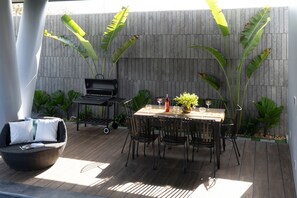 Een terras buiten met een ronde tafel, stoelen, een barbecue en grote planten.