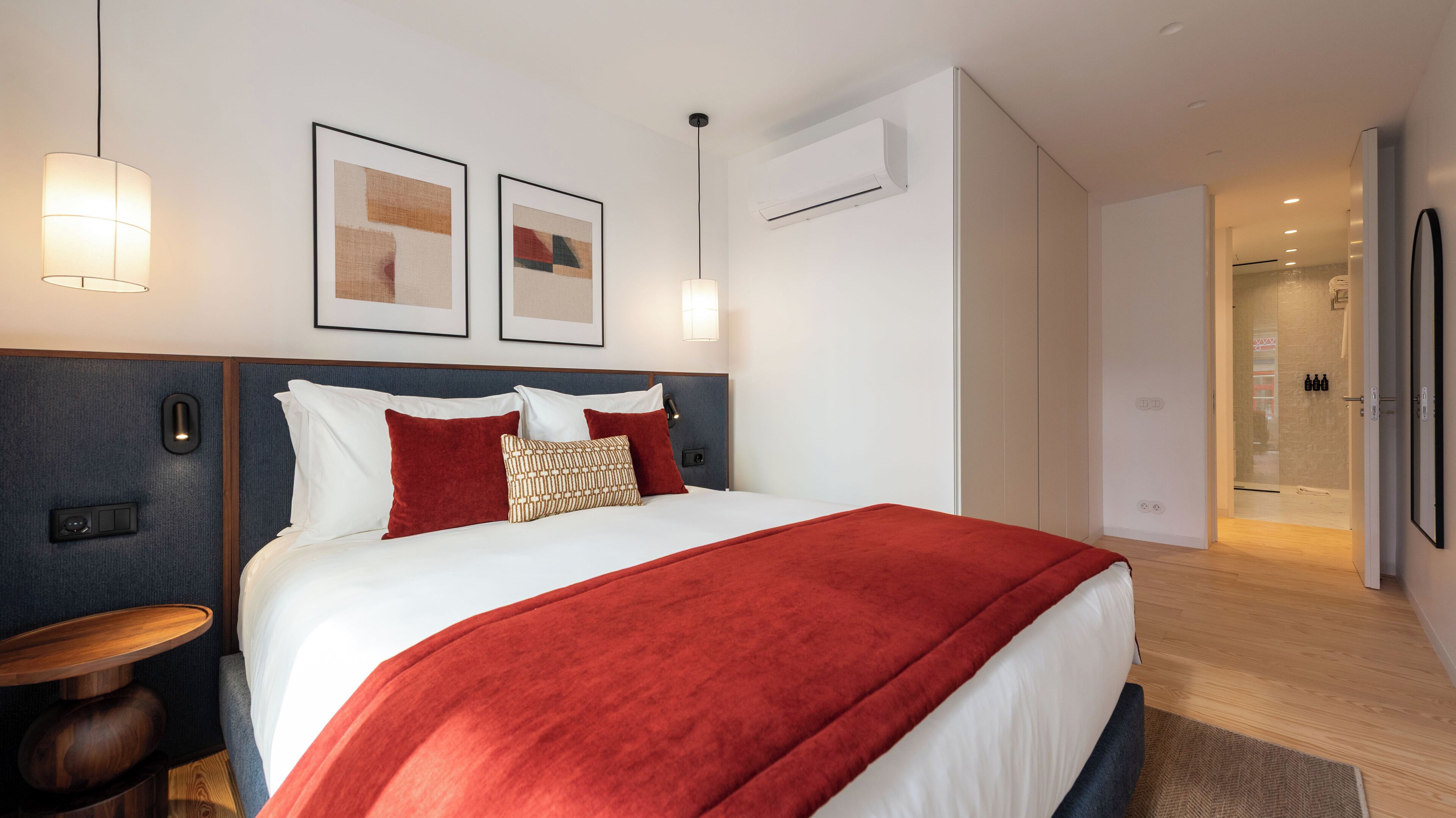 A modern hotel room with a large bed, a bedside table, a nightstand, a wall-mounted air conditioning unit, and a door leading to a shower area.