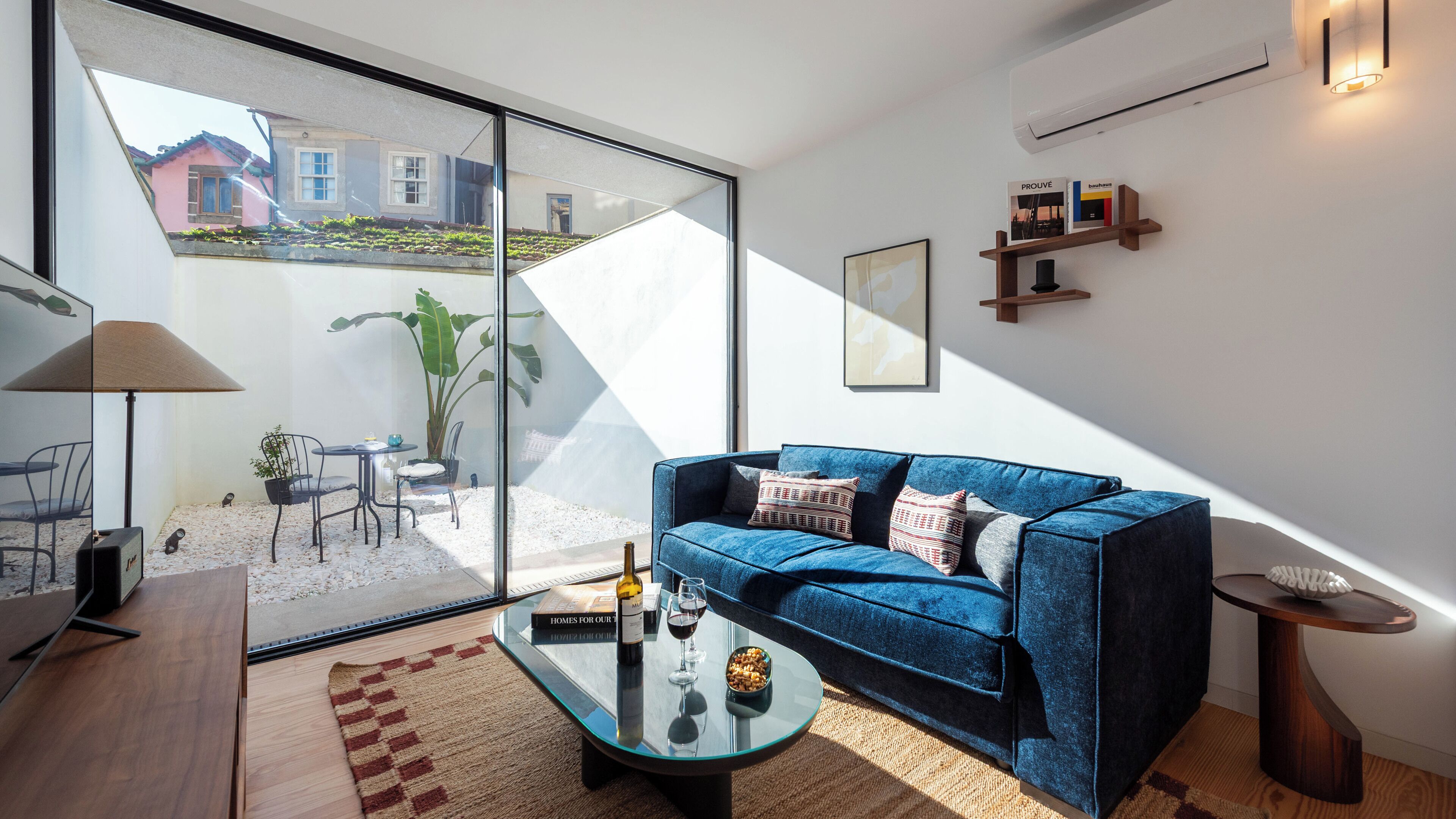 A modern living room with a blue sofa, a glass coffee table, and a view of a garden through large windows.