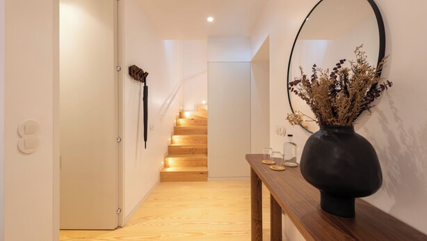 Un couloir avec un escalier en bois, un vase contenant des fleurs séchées et un miroir accroché au mur.