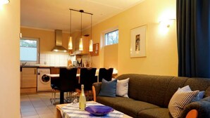 A cozy living room with a sofa, a coffee table, and a dining area with chairs. There is a kitchen in the background with a window. - Vacation home in Tossens - relaxation for the whole family. (Butjadingen)