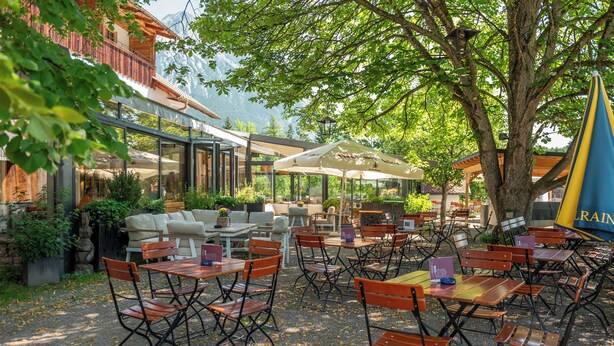 An outdoor seating area with tables and chairs, surrounded by greenery and a building with a balcony. - Sonnenplateau Cabins by amii (Wildermieming)