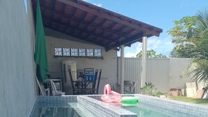 A swimming pool with tiled edges and a covered area with chairs and a table. - Lar Jampa Praias Sul (Conde)