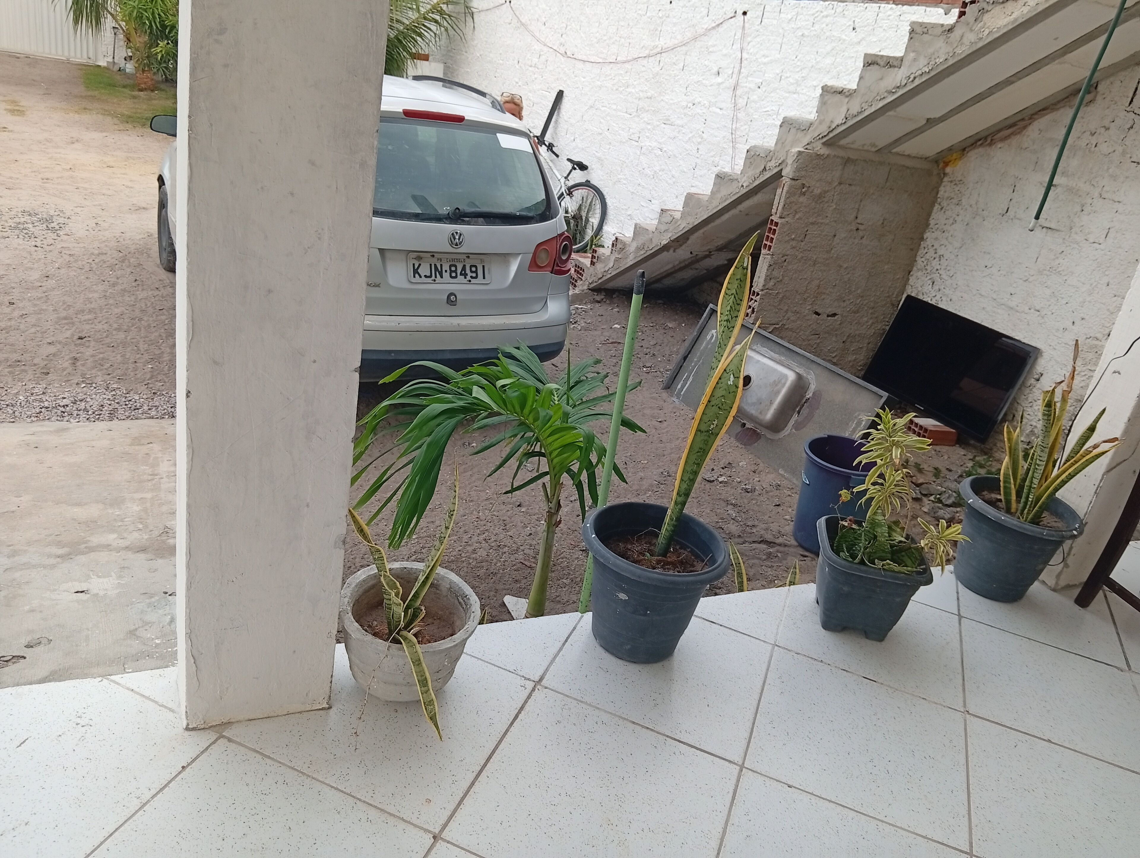 A balcony with potted plants, a white car parked outside, and a TV on a wall.