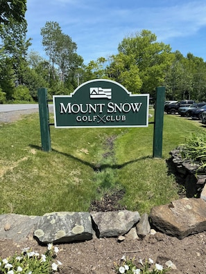 A sign for Mount Snow Golf Club with a clear sky and parked cars in the background.