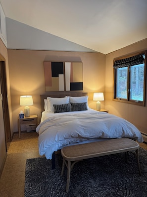 A bedroom with a bed, two bedside tables, a window with a view of trees, and a wall-mounted abstract artwork.