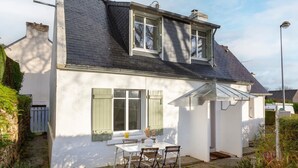 Une maison à deux étages, avec une façade blanche, un toit en pente et une petite terrasse équipée d’une table et de chaises.