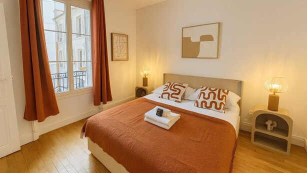 Ein ordentlich bezogenes Bett mit einer braunen Decke, zwei Kissen mit geometrischem Muster und einem Nachttisch mit Lampe.