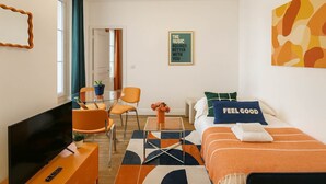Ein modernes Hotelzimmer mit einem Glastisch, orangefarbenen Sesseln, einem Bett mit orangefarbener Bettwäsche, einem Fernseher und einem gerahmten Motivationsspruch an der Wand.