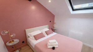 Un dormitorio con cama, almohadas, una mesita de noche, una lámpara y una ventana.