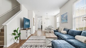 A modern living room with a blue sofa, a coffee table, and a TV. There is a staircase on the left and a dining area in the background. - Holy City Hideaway | Minutes from Everything (North Charleston)