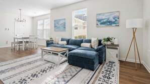 A modern living room with a blue sectional sofa, a wooden coffee table, and a patterned rug. There is a dining area with a white table and chairs, and a painting on the wall. - Holy City Hideaway | Minutes from Everything (North Charleston)