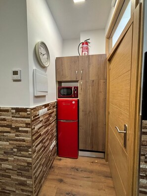 A compact kitchen with a red refrigerator, wooden cabinets, a microwave, a clock, and a fire extinguisher on the wall.