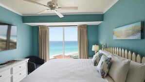 Une chambre à coucher comprenant un lit, un ventilateur de plafond, une télévision, un bureau, une chaise, une fenêtre avec des rideaux et un tableau.