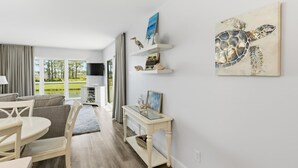 Una sala de estar moderna con una mesa de centro de vidrio, un sofá y un televisor. Hay objetos decorativos en estanterías y un cuadro de una tortuga marina.