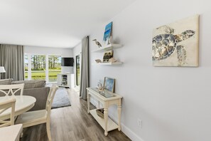 Ein modernes Wohnzimmer mit einem Couchtisch aus Glas, einer Couch und einem Fernseher. Auf Regalen stehen Dekoartikel und es hängt ein Gemälde von einer Meeresschildkröte.