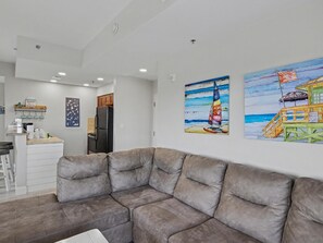 A modern living room with a sectional sofa, a kitchen area with wooden cabinets, and two beach-themed paintings on the wall. - 1BR Luau Condo on 7th Floor at Sandestin Golf & Beach Resort (Miramar Beach)