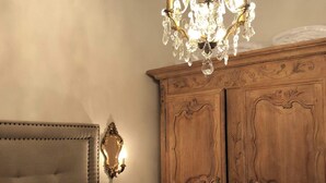 Quarto com cabeceira estofada, guarda-roupa de madeira com detalhes entalhados e um lustre de cristal.