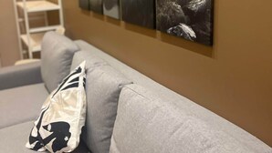 Un divano grigio con un cuscino decorato, una scaffalatura bianca e una serie di dipinti con paesaggi alle pareti.