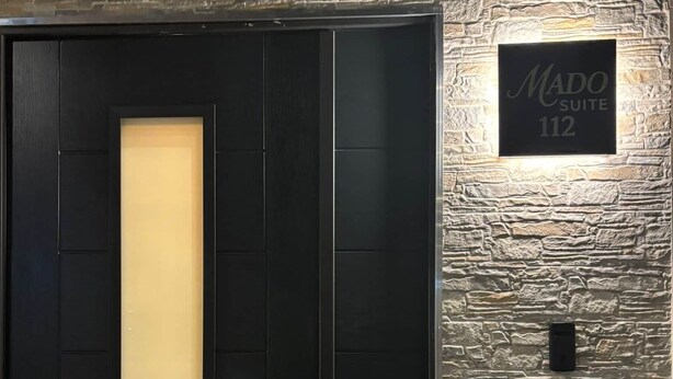 Una porta nera con un pannello verticale in vetro satinato, una serratura e un campanello. Sopra la porta, un cartello con la scritta 'Mado Suite 112'.