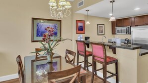 A dining area with a glass table and wooden chairs, a chandelier, and a kitchen with a microwave and refrigerator. - Stylish 1BR condo in The Grand at Sandestin Golf & Beach Resort! (Miramar Beach)