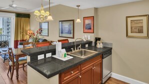 A modern kitchen with a granite countertop, stainless steel sink, and wooden cabinets. There are framed artworks on the walls and a dining area with a table and chairs. - Stylish 1BR condo in The Grand at Sandestin Golf & Beach Resort! (Miramar Beach)