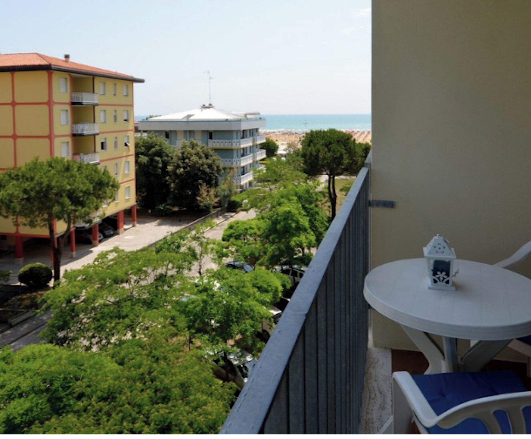 Un balcón con vista a la playa, un edificio de fachada roja y amarilla, y una mesa blanca con un pequeño adorno.