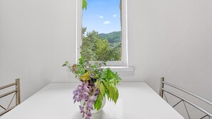 Ein weißer Tisch mit einer Metallumrandung, ein Fenster mit Blick auf grüne Hügel und eine Vase mit Blumen.