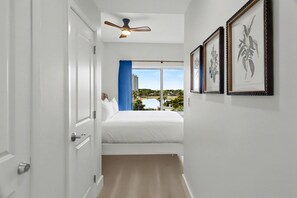 Ein Hotelzimmer mit Bett, Deckenventilator, Fenster mit Blick auf Gebäude und Grünflächen sowie gerahmten Pflanzenbildern an der Wand.