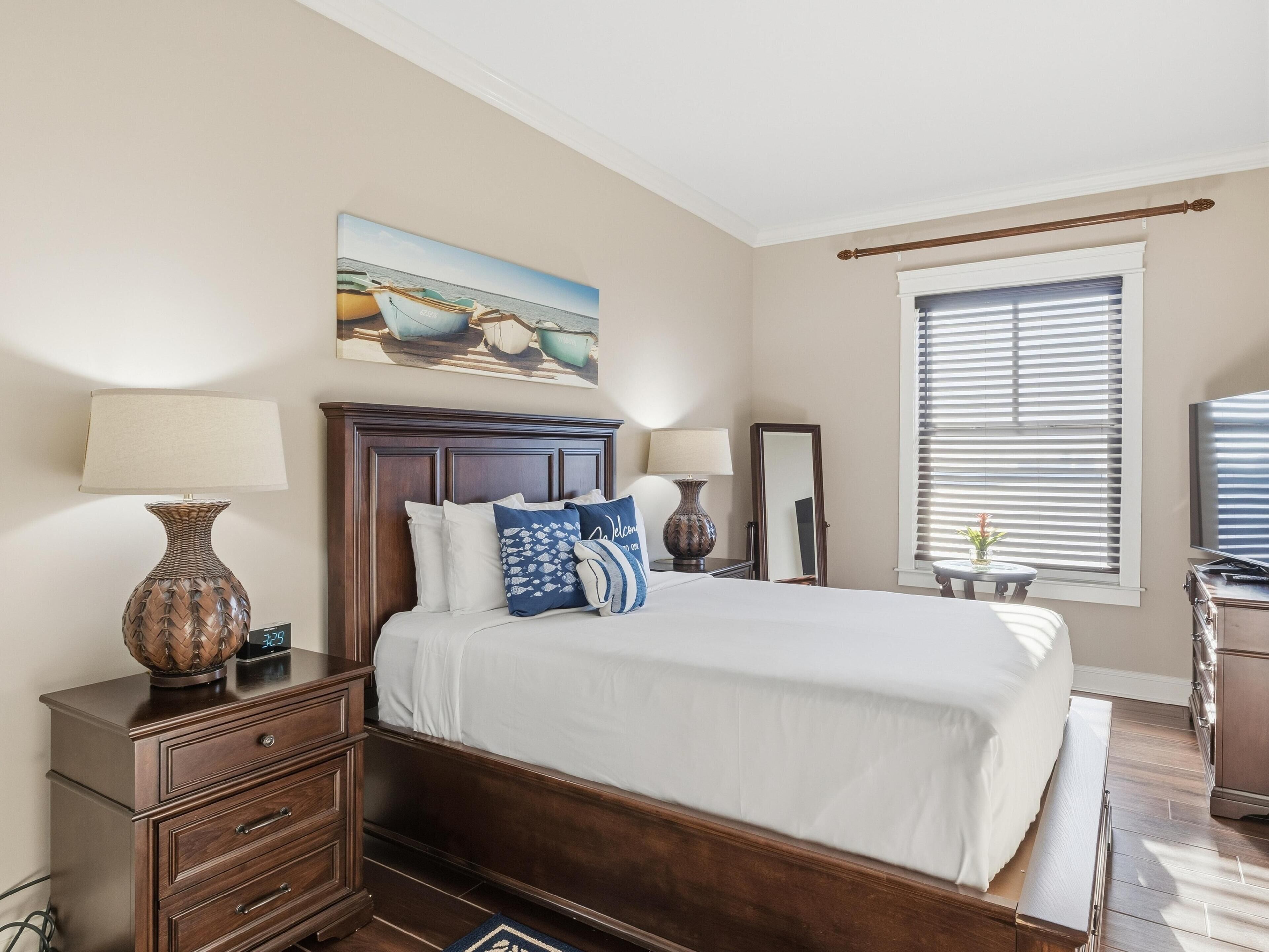A bedroom with a large bed, two bedside tables, a nightstand, a mirror, a window with blinds, and a painting on the wall.