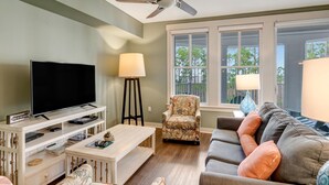 A living room with a flat-screen TV, a ceiling fan, a sofa, a patterned armchair, and a window with a view of trees. - 2BR Bahia Suite at Golf & Beach Resort (Miramar Beach)