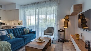 A modern living room with a blue sofa, a wooden coffee table, and a chair. There is a kitchen in the background with white cabinets and a sink. - Starlet 1-BR Apt. & Pool in Larnaca (Larnaca)