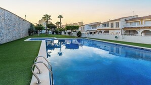 A swimming pool with a stone wall on one side, surrounded by well-maintained grass and palm trees. - Sun-Kissed 3BR Townhouse by Pool, Near Carabassi Beach (Gran Alacant)