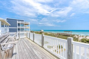 Eine Holzterrasse mit weißer Brüstung, die auf einen Strand und ein Gebäude mit mehreren Balkonen blickt.
