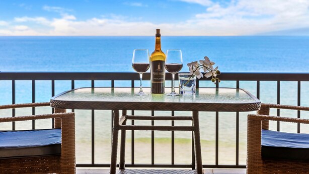 A table with a bottle of wine, two glasses, and a small glass of water on a balcony with a view of the ocean. - 8th Floor Oceanfront | 180° Sunset Views (Honokowai)