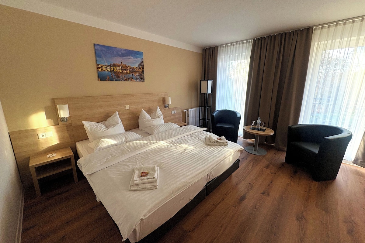 A hotel room with a large bed, a chair, a small table, and a painting on the wall.