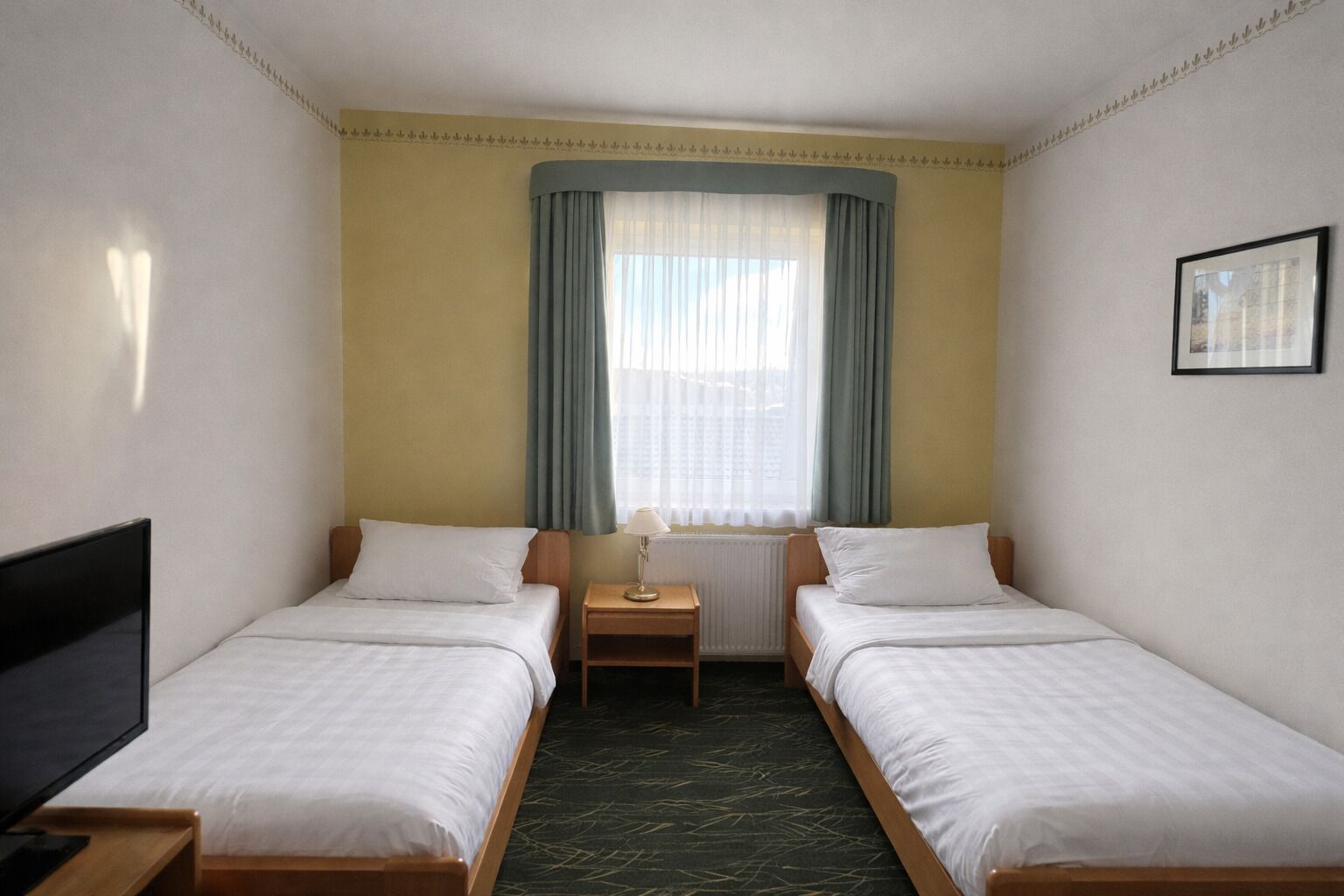 Une chambre d’hôtel avec deux lits simples, une petite table, une lampe, une télévision et une fenêtre avec des rideaux.